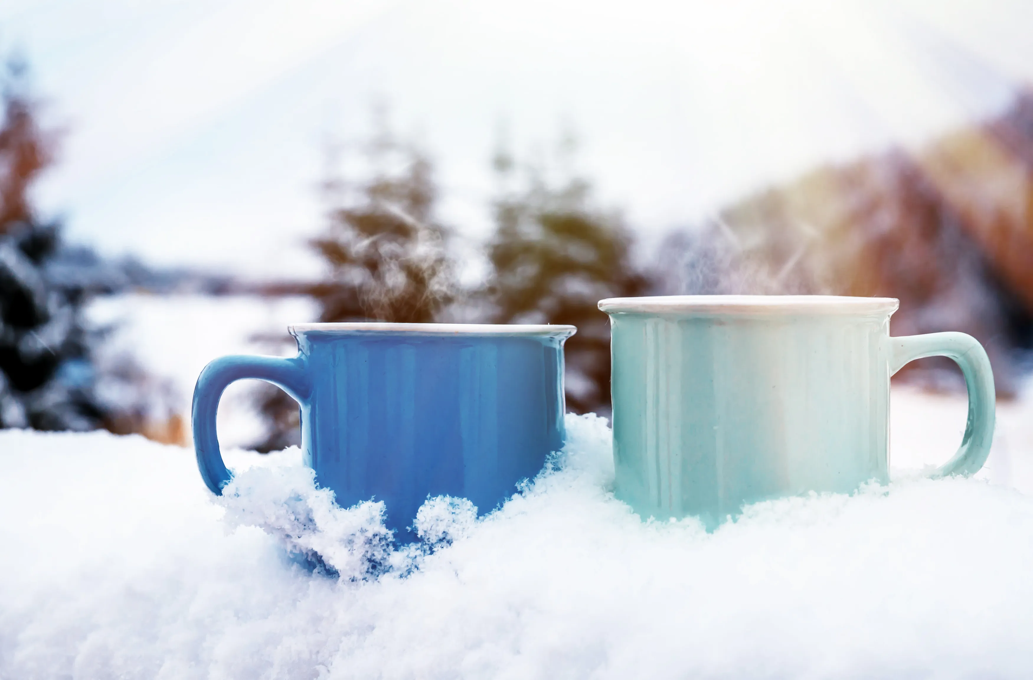 Two steaming mugs of coffee rest on an outdoor bannister. Snow is packed around the mugs and the heat from their coffee is melting the snow.