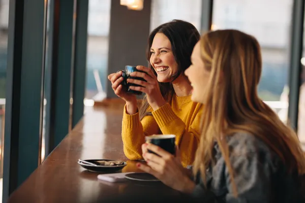 Two women in sweaters chat happily at their local coffee-shop. They are sitting at a bar height table next to a bright and sunny window.