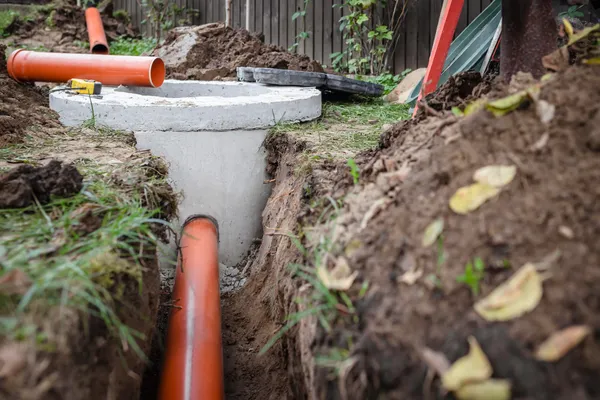 Workers dig a trench around the orange pipes of a septic tank so that they can repair them. Pieces of new pipe can be seen laying next to the trench.