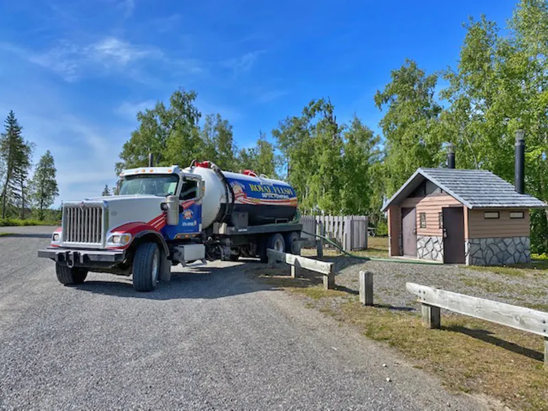A Royal Flush pumping truck backed up to an outhouse at a local park. The pumping hose can be seen snaking into the outhouse.
